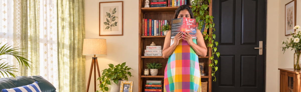 Curator: a woman in a checked cotton dress holds up a book beside a bookshelf in a sunlit living room