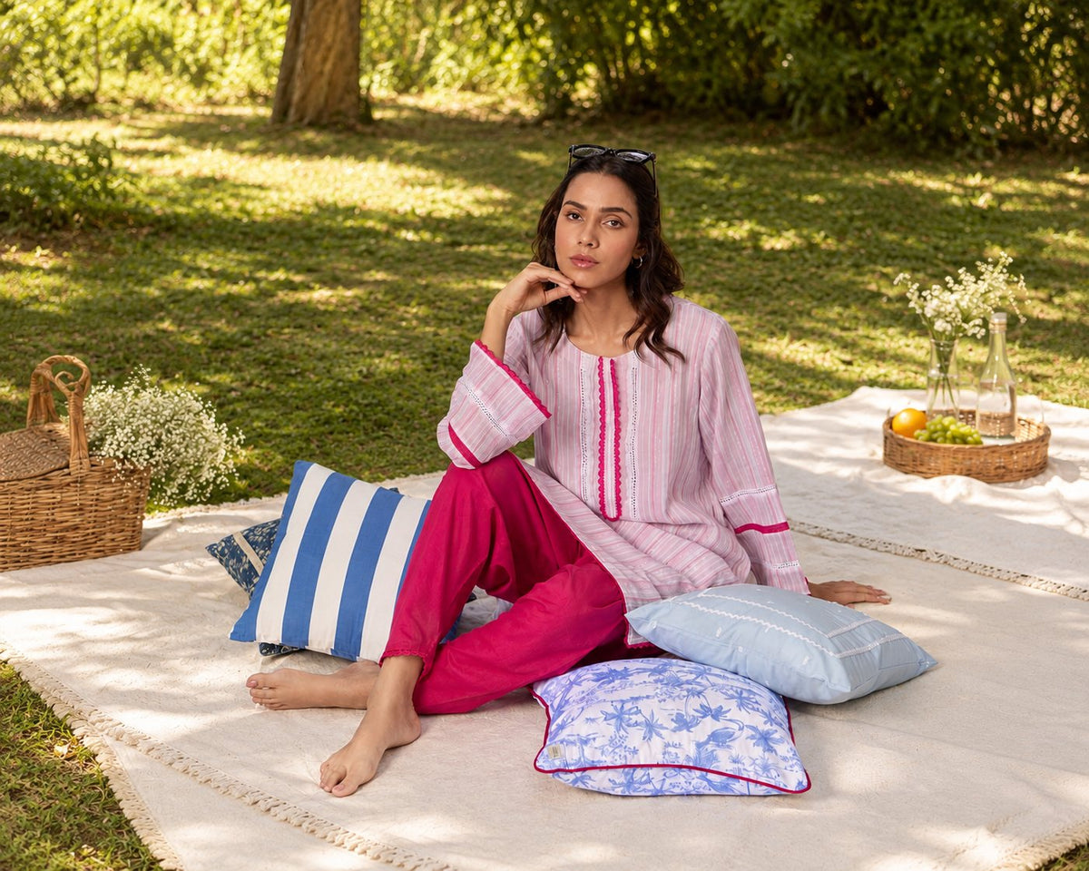 A woman in a pink kurta sits on a picnic rug with striped and floral cushions in a garden